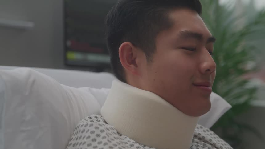 A male patient with a neck brace reclines in a hospital bed with a faint smile