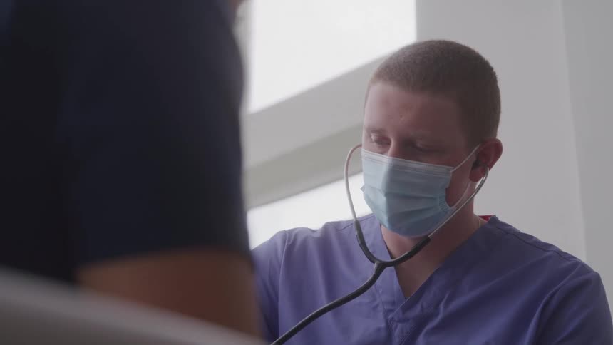The video shows a male doctor wearing a mask and scrubs using a stethoscope to listen to a patients chest the patient is not clearly visible in the foreground of the shot