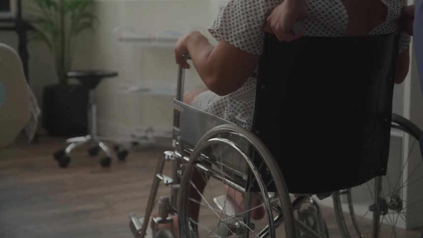 A female patient in a hospital gown is being pushed in a wheelchair by a healthcare worker