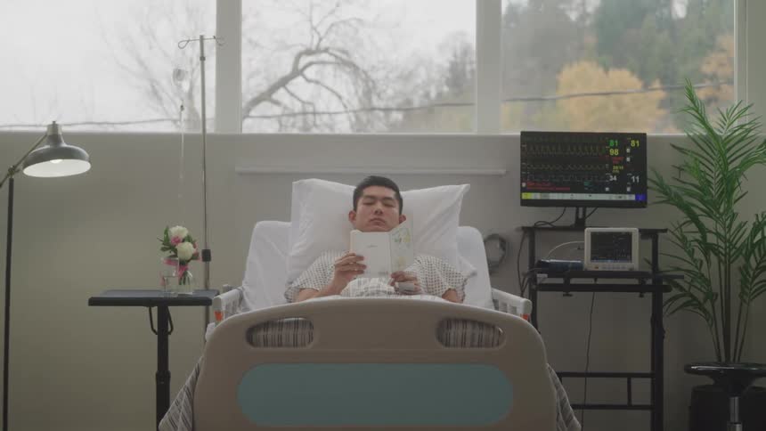 A young man is lying in a hospital bed propped up with pillows and reading a book or magazine a heart rate monitor is visible next to the bed along with an iv pole and a potted plant
