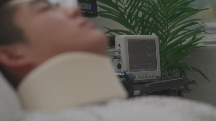 The video shows a closeup of a patient lying in a hospital bed with a heart rate monitor beside them the monitor displays the patients vitals and a plant is visible in the background
