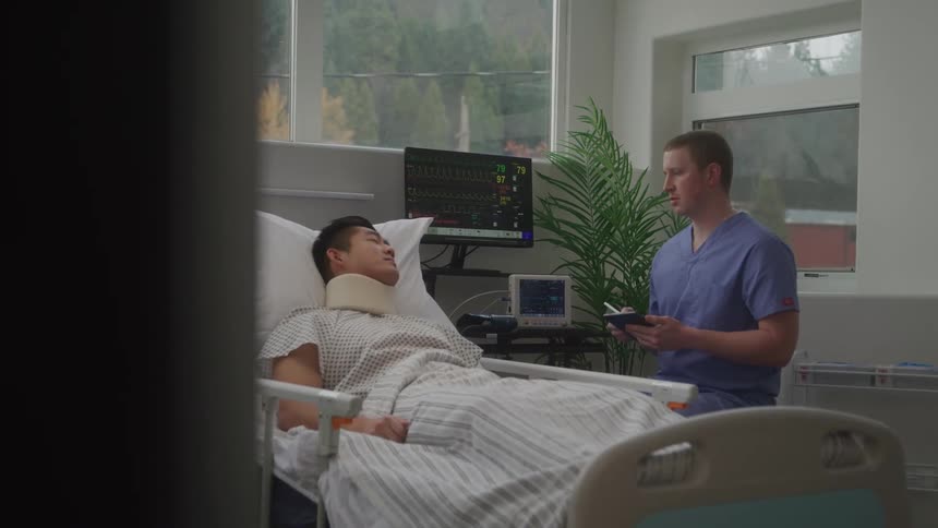 A male patient with a neck brace rests in a hospital bed while a male nurse attentively takes notes