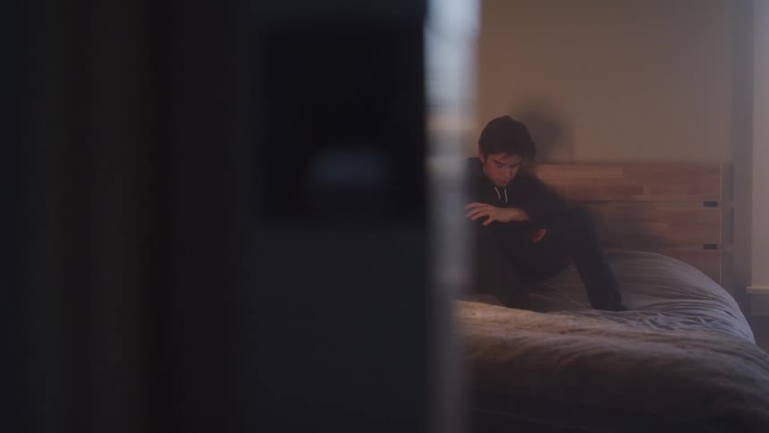 A young person is sitting on a bed in a dimly lit room appearing to be deep in thought the shot is partially obscured by a dark foreground object framing the subject in the background