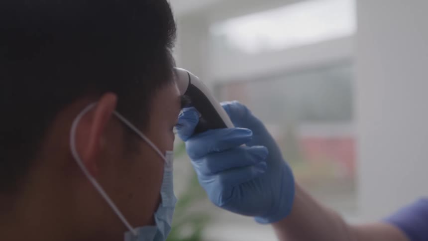 A doctor wearing a blue glove and a face mask holds a thermometer to the forehead of a male patient who is also wearing a face mask