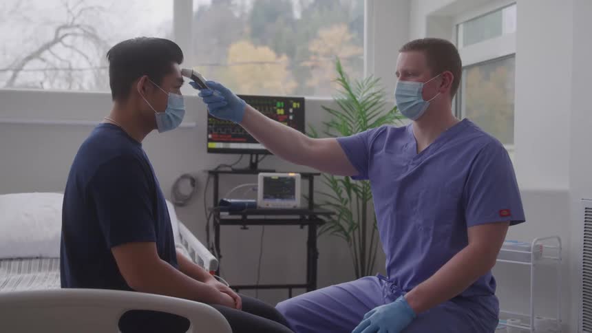 A nurse checks the temperature of a patient in a hospital room with both men wearing nose masks