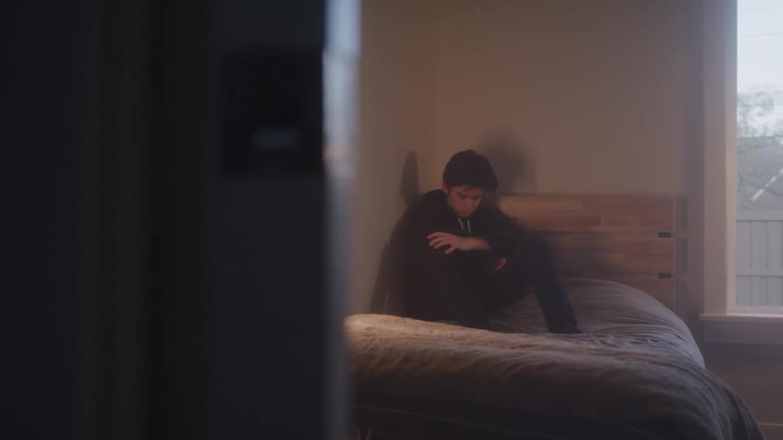 The video shows a person sitting on a bed in a dark room with their head in their hands the shot is framed from a doorway showing a window on the right side of the room