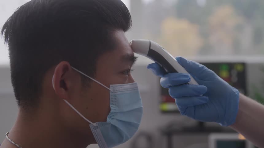 A masked male healthcare worker faces a thermometer likely during a routine check in a clinical setting