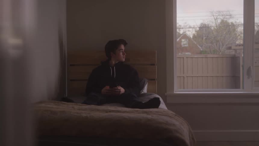 A wide cinematic shot of a young person sitting alone on a bed engrossed in their phone with natural light coming in from a large window to their side