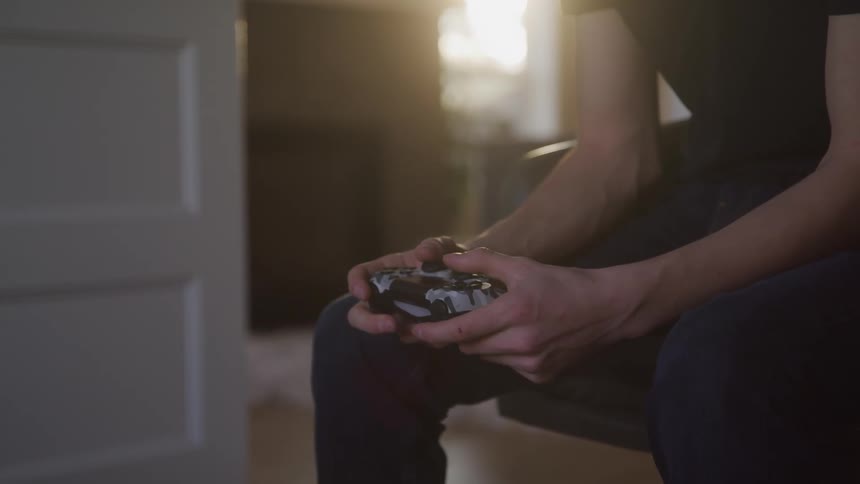 A male sits indoors gripping a game controller with both hands in soft light
