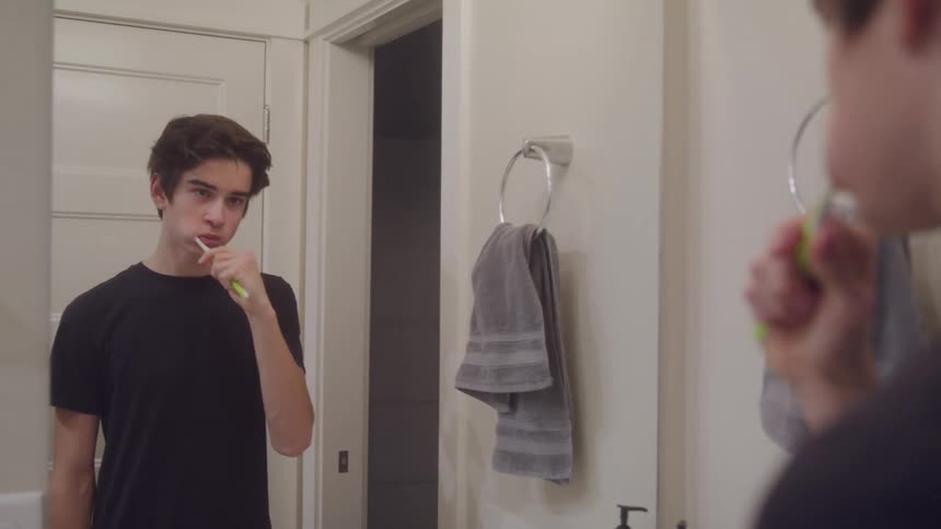 A teenage boy is brushing his teeth in front of a bathroom mirror