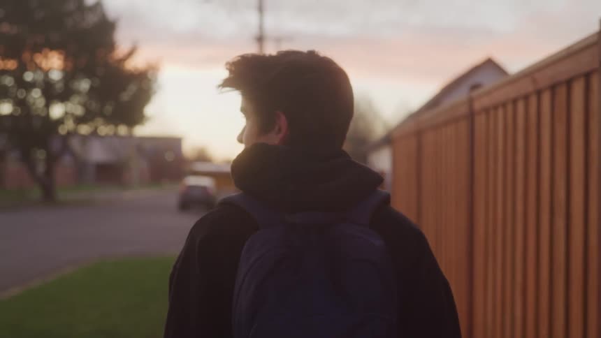 The video shows a person with a blue backpack walking down a residential sidewalk at sunset they walk with a purposeful stride passing by a wooden fence and a large tree as the day ends