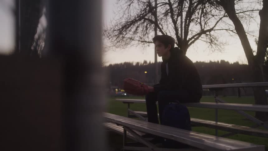 A teenage boy with dark hair and a black hoodie sits alone on a park bench at dusk holding a baseball glove in his lap