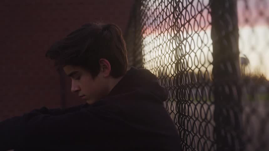 A teenage boy sits on the floor beside a wire mesh fence at sunset