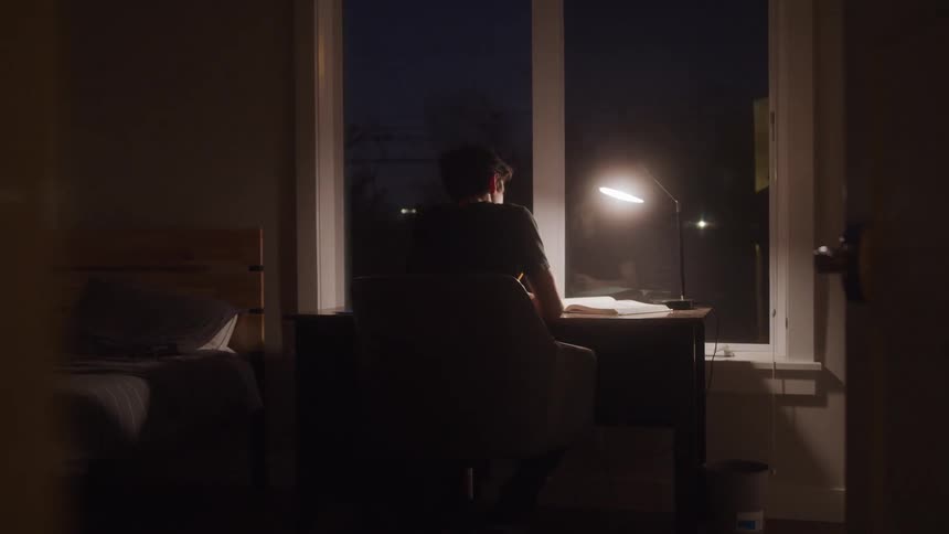 A man in his room studying reading a book at his desk