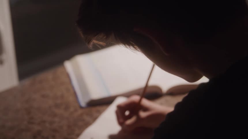 A person with a dark shirt and a book in front of them is writing with a pencil the shot is a closeup from behind focusing on their hand and the notebook