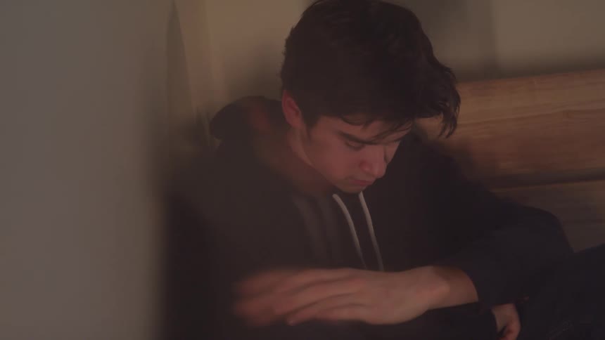 A young man with dark hair is sitting down looking down and to the side he is wearing a black hoodie and appears to be in a thoughtful or melancholic state