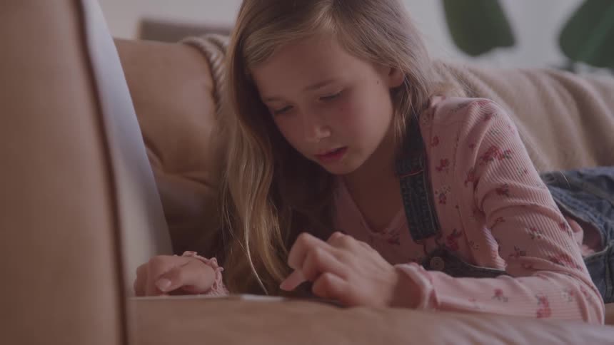 The video shows a lowangle shot of a young girl lying on a couch intently using a tablet she has long blonde hair and is wearing a pink shirt and denim overalls