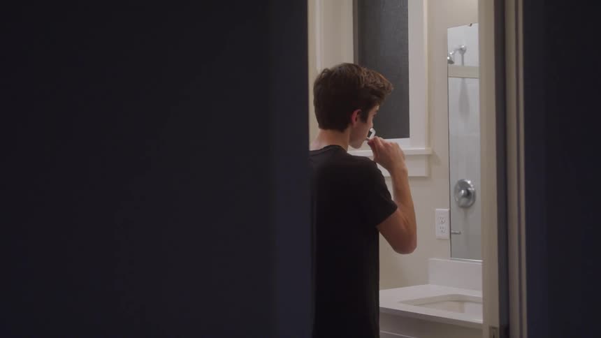 The video shows a young man brushing his teeth in a bathroom while looking into a mirror the shot is framed through a doorway with the mans reflection visible as he continues his hygiene routine