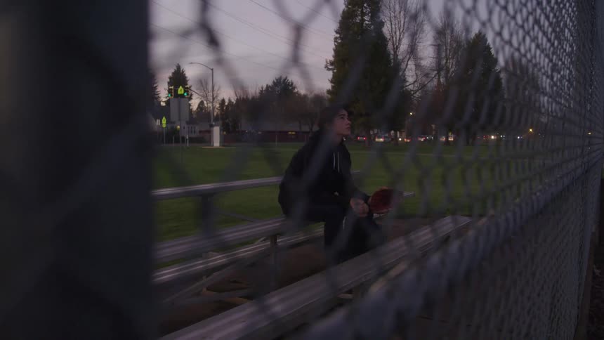A young man sits alone on a bench at a sports field holding a baseball glove the shot is taken from behind a chainlink fence and houses and trees can be seen in the background