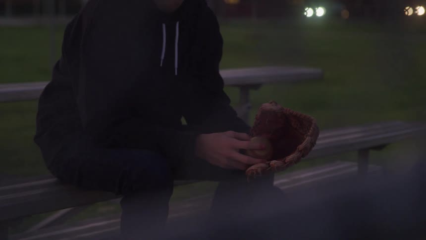 A person sits on a park bench at night wearing a black hoodie and black pants playing with a baseball in a glove