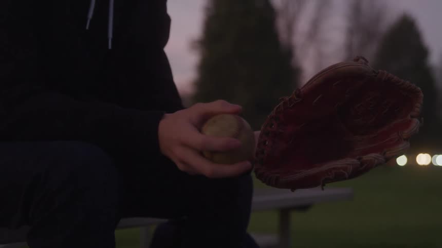 A man sitting near a field tossing a baseball in his hand wearing a baseball glove