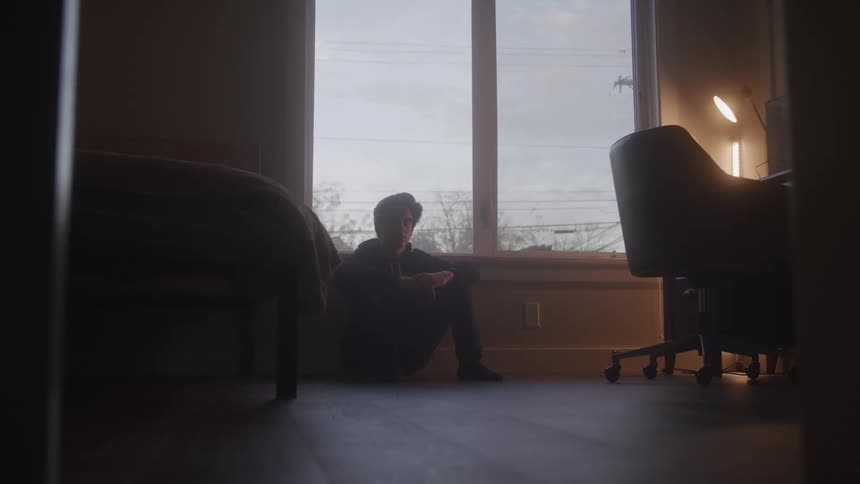 Person sits alone by a window in a dimly lit room looking sad