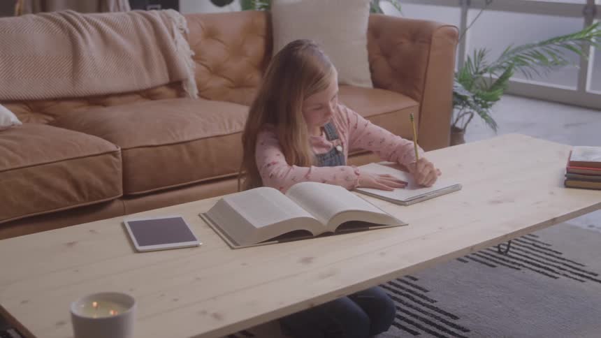 The video shows a young girl with long blonde hair kneeling at a wooden table in front of a brown couch she is writing in a notebook and there is an open book and a tablet on the table with her