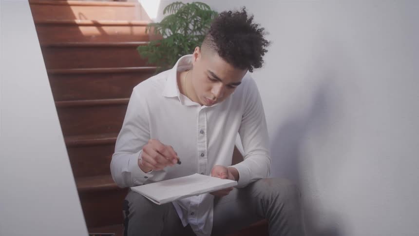 A young man in a white shirt sits on stairs focused on sketching in a notebook