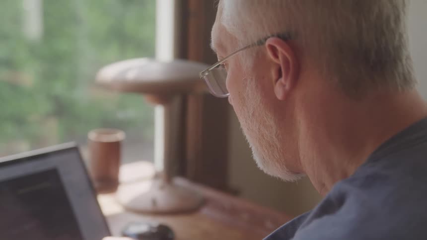 Elderly man works on laptop near a window in natural light