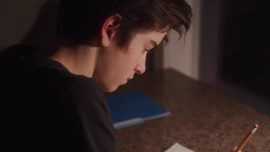 A young male concentrates on writing under warm lighting at a countertop
