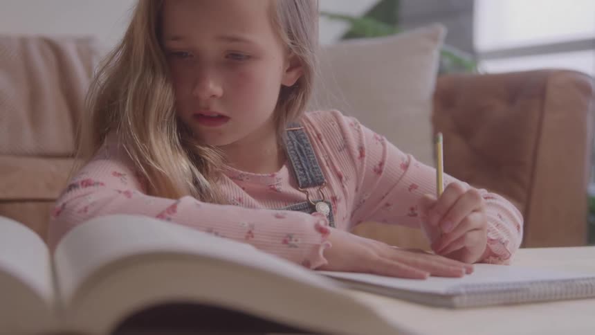 A young girl with long hair leans on a table while writing in a notebook