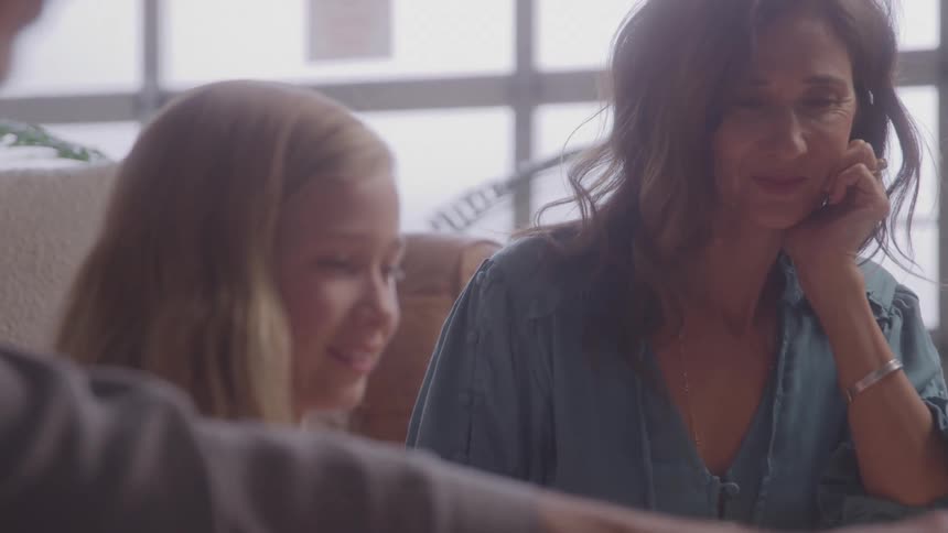 The video shows a woman with dark hair sitting on a couch with a young girl the woman is smiling and looking at the girl who is also smiling and a man is partially visible in the foreground