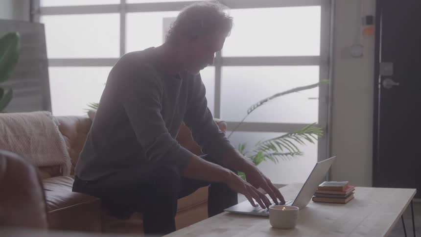 A man with grey hair sits on a brown couch and works on a laptop that is resting on a coffee table in a brightly lit room