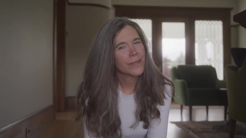 A woman with long hair in a white top smiles softly while sitting in a warmly lit living room