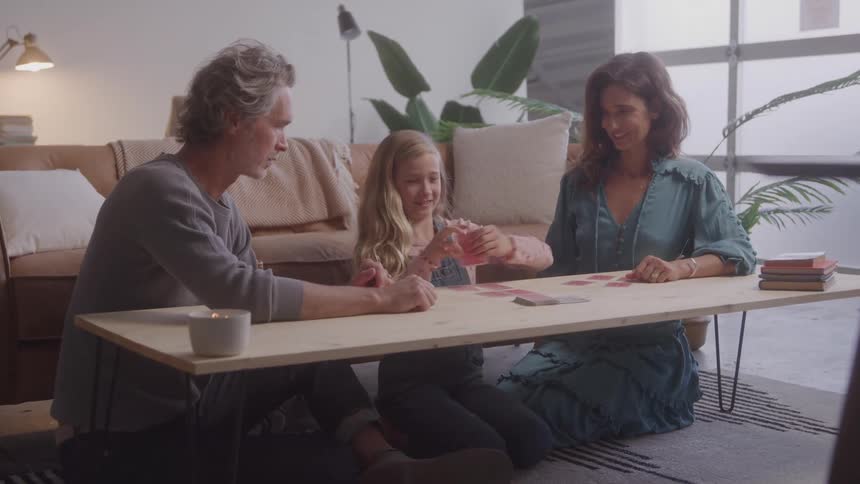 A mother and father sit on the floor and play a card game with their young daughter and they all smile as they look down at the cards on the low wooden table
