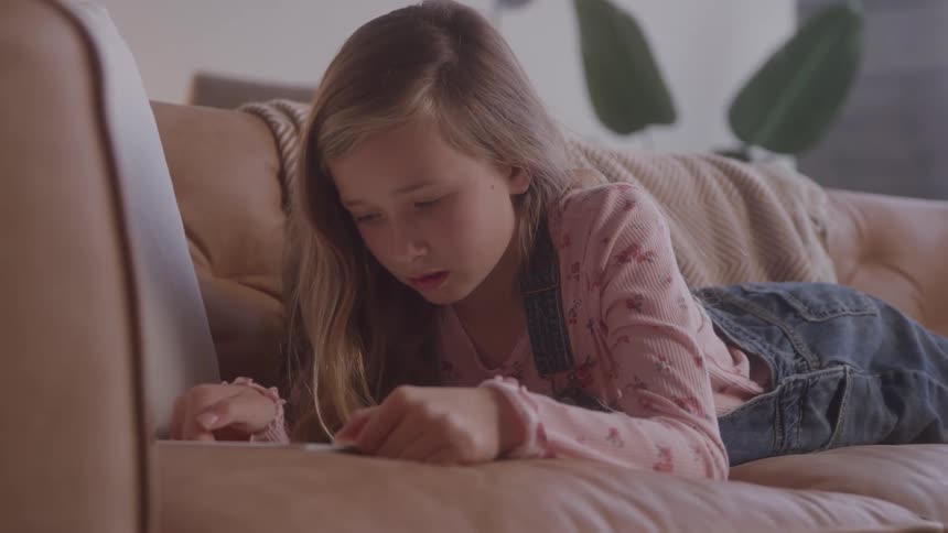A young girl lies on a couch focused on a phone she holds in both hands