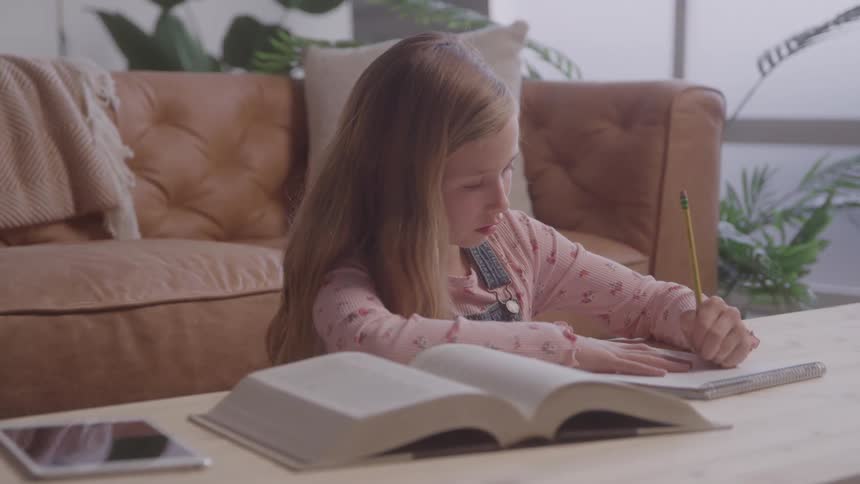 Little girl in living room sitting writing and studying with an open book