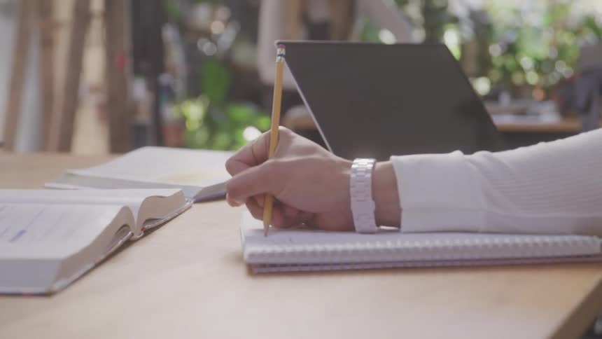 Person writes in a notebook with a pencil next to an open book and a laptop on a wooden table