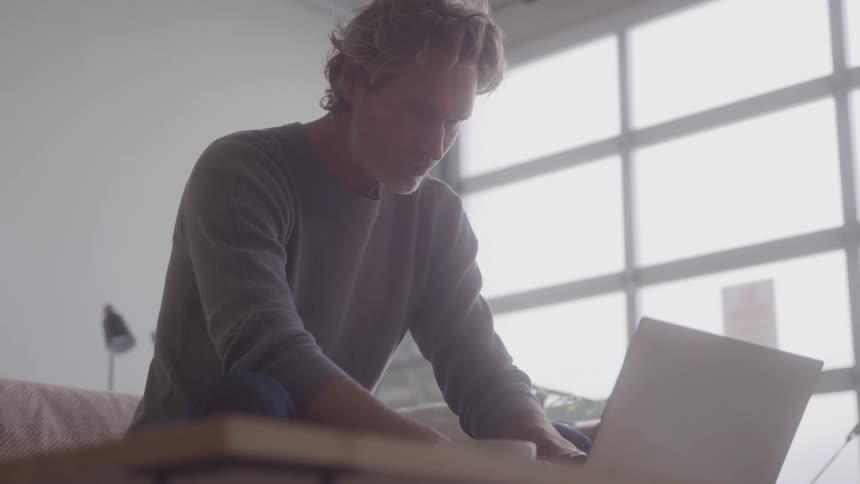 A man leans over a table working on a laptop in a bright office