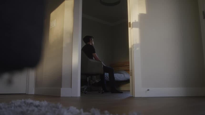 A young man sits alone in a dimly lit bedroom at sunset facing away with a contemplative posture