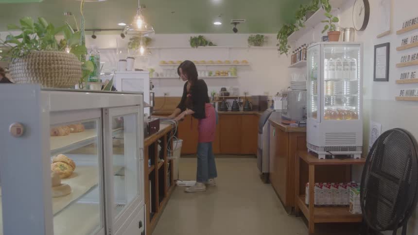 A barista in a black shirt and jeans with her hair tied back is wiping down a counter behind a coffee shop display case
