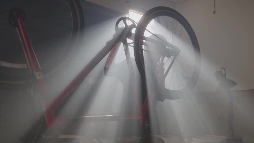 A bicycle mechanic works on a red bicycle which is upside down on a repair stand in a dusty garage as rays of sunlight stream through the garage door illuminating the particulate matter in the air