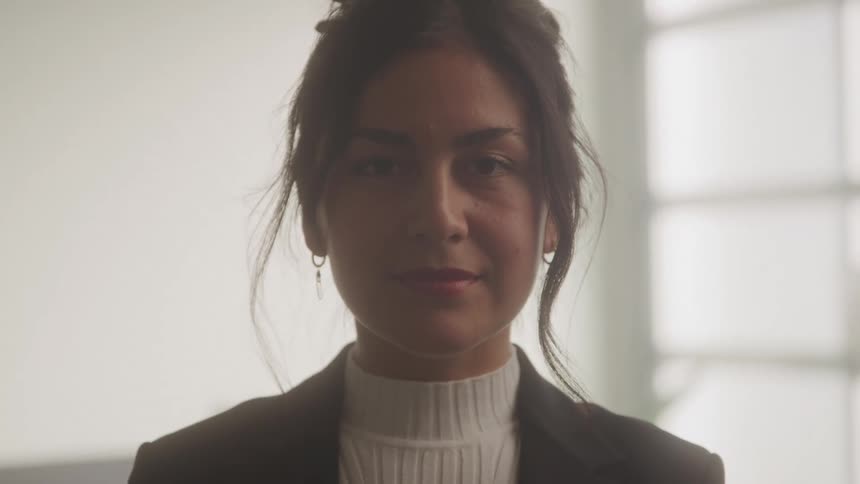 A young woman wearing a turtleneck shirt a suit and earrings stands inside an office at midday with a smile on her face
