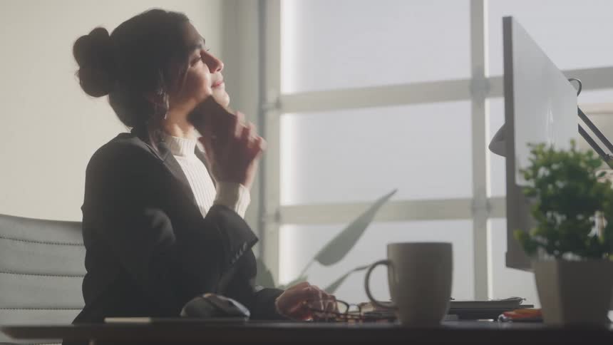 A young woman wearing a turtleneck shirt and a suit sits in front of a desk with a computer monitor in an office answering a call on her phone she leans back with a smile while talking with a flower plant and a mug on the side of the desk at midday