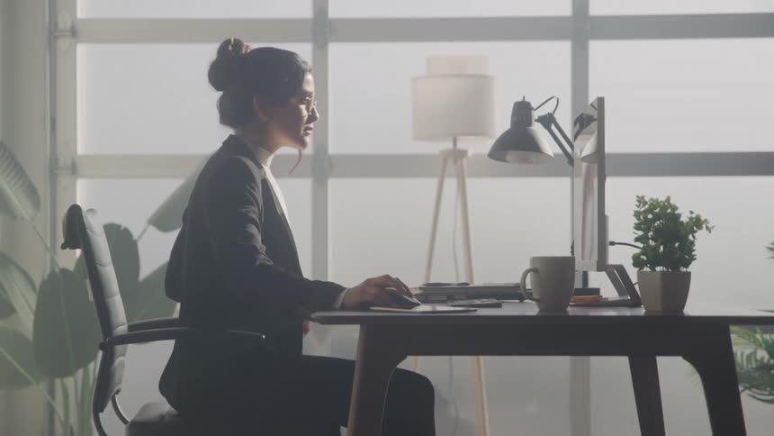 A professionallooking woman with her hair in a bun is sitting at a desk in an office working on a computer she is seen from the side wearing a black blazer and glasses and is partially silhouetted against a large window