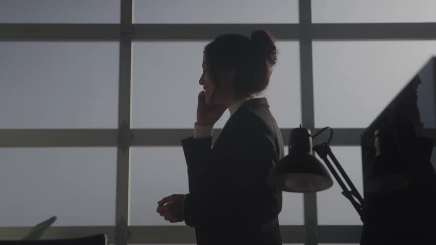 A young woman wearing a black suit stands inside her office at sunset talking on the phone