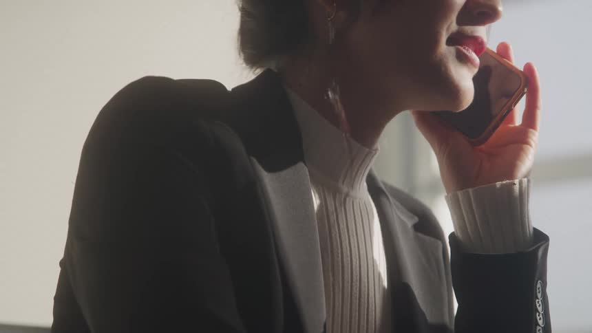 A young woman wearing a suit and a turtleneck shirt sits in an office answering a call on her mobile device at midday