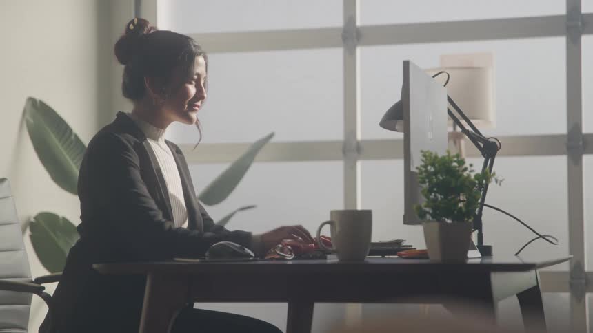 A person is working at a computer in a brightly lit office their back to the window they are wearing a black blazer and have their hair tied back in a bun