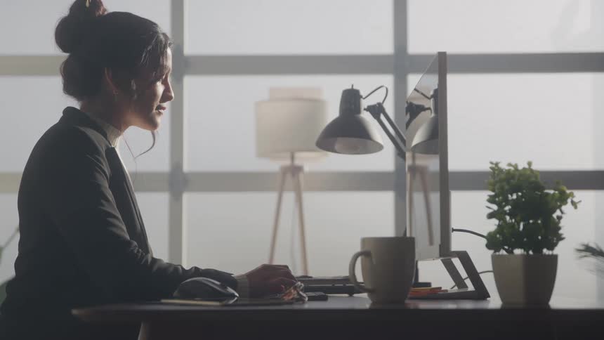 A young woman wearing a suit types on her computer in her office at midday with a reading lamp mug and potted plant on the desk