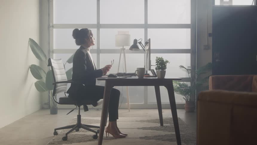 A young woman in a black blazer and heels walks into a modern bright office and sits down at a desk she takes out her earbuds and begins to work on a laptop